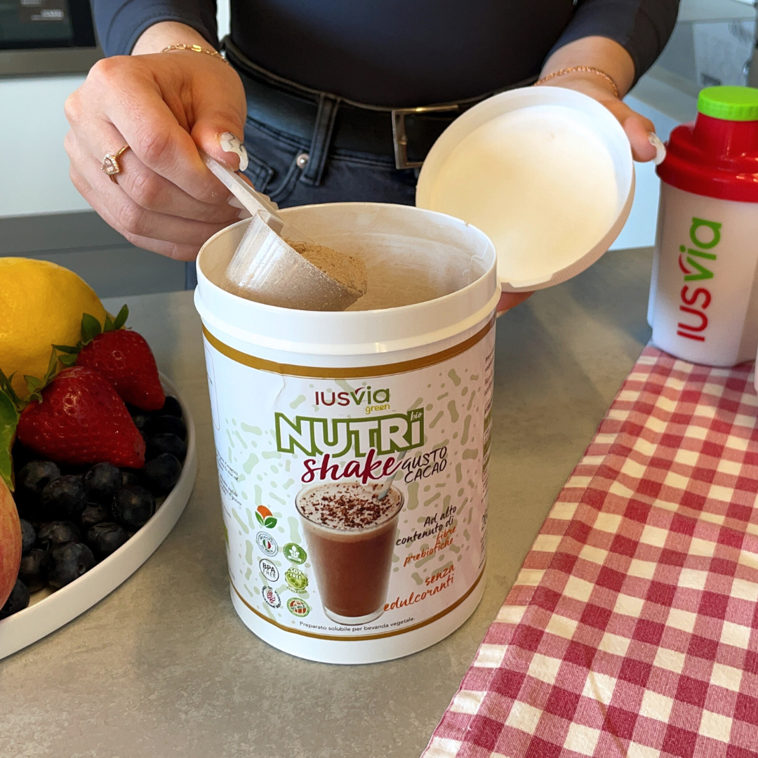Immagine con il prodotto Green Nutri Shake Al Gusto Cacao. Si vede un persona che sta prendendo un misurino di prodotto. Sullo sfondo una tovaglia a quadri rossa e bianca, lo shake IUSVIA, e una fruttiera con frutta fresca.