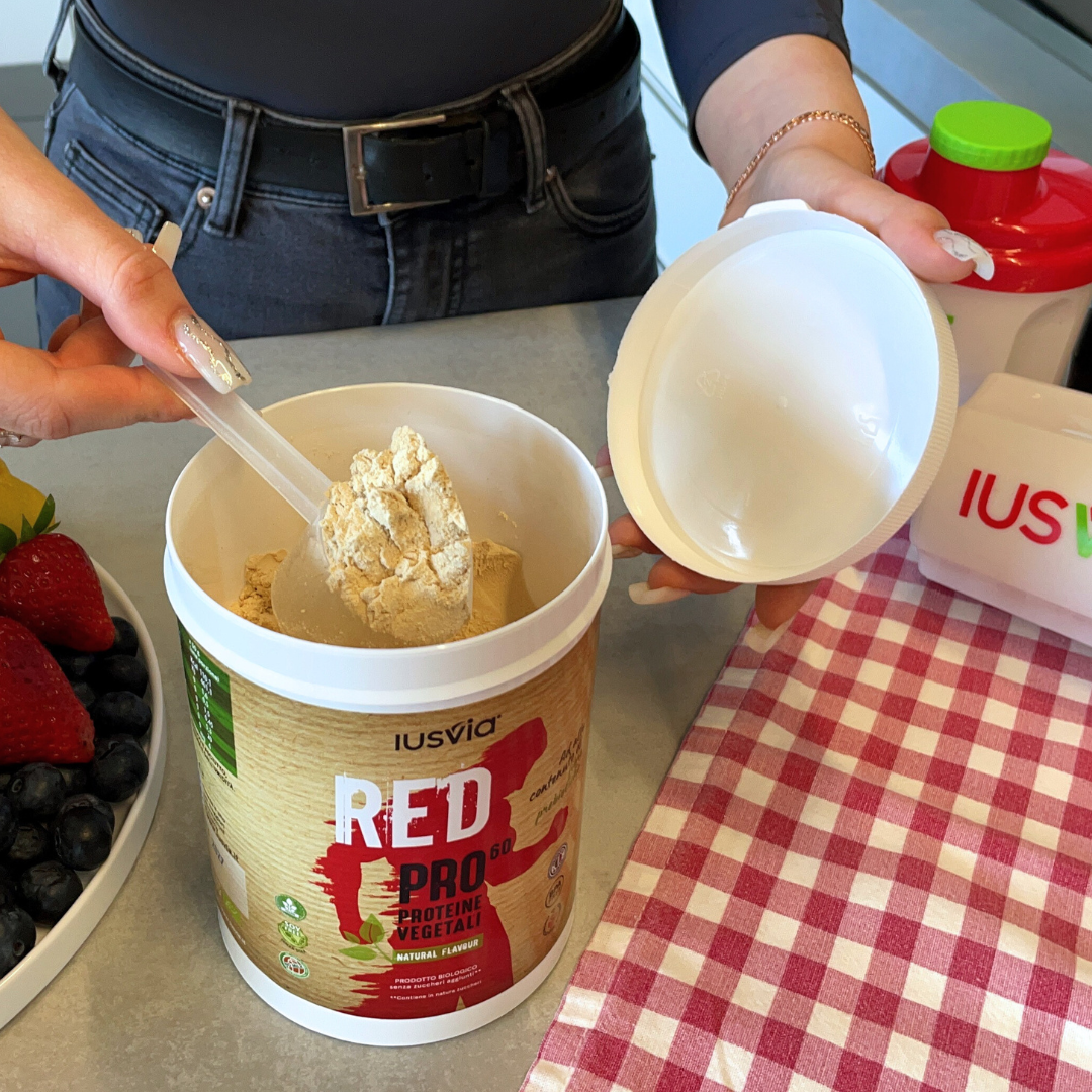 Prodotto RED PRO 60 Proteine Vegetali Natural Flavour. Si vede una persona che prende un misurino di prodotto, nello sfondo una tovaglia a quadri rossi e bianchi, uno shake e una fruttiera con frutta fresca.
