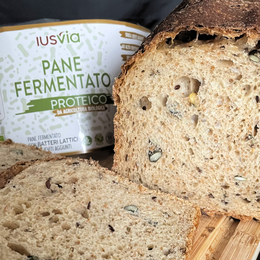 Foto prodotto Pane Fermentato Proteico da Agricoltura Biologica. Si vede nella foto il pane proteico tagliato a fette sopra un tagliere di legno. Sullo sfondo l'etichetta del prodotto.